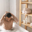 wet-young-muscular-man-washing-his-hair-while-having-bath-with-foam-by-shelves-with-self-love-items