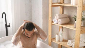 wet-young-muscular-man-washing-his-hair-while-having-bath-with-foam-by-shelves-with-self-love-items